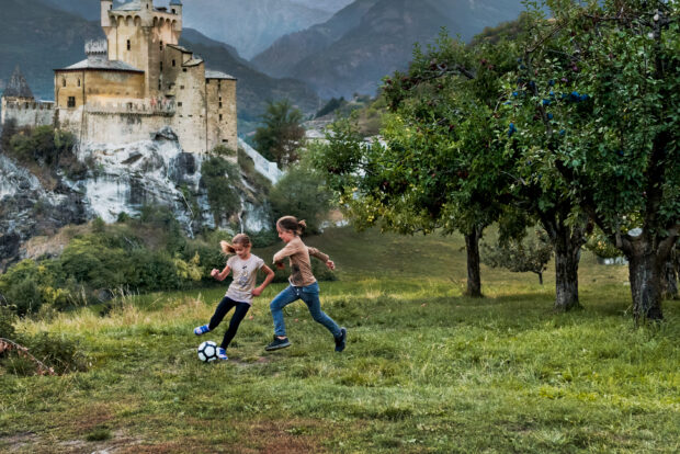 Children playing soccer in an orchard with a castle in the background.